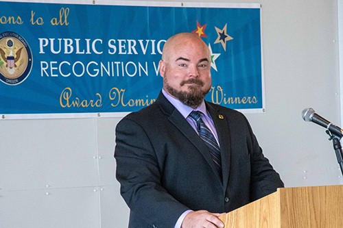 Photo of Brock Young in business attire standing at a podium.  Behind him is a banner with the text "Purblic Service Recognition Week - Award Nominee Winners"